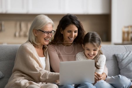 Photo for Happy kid girl, mom and granny having fun at laptop, resting on couch together, using computer for Internet communication, watching movie on media service, laughing - Royalty Free Image