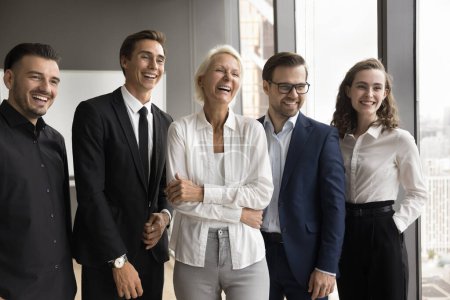 Photo for Joyful younger colleagues and happy elder team leader woman standing together, smiling, laughing, looking away, celebrating corporate achievement, teamwork success. Casual business portrait - Royalty Free Image