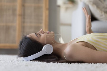 Photo for Peaceful positive pretty teenager girl in big wireless earphones lying on back at home, resting on soft carpeted floor, listening to music, singing song, enjoying leisure, rhythm - Royalty Free Image