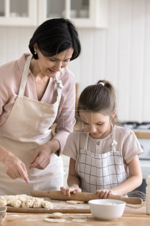 Photo for Vertical shot mature 60s woman teach to cook her granddaughter. Little girl flattening dough using rolling-pin, enjoy cookery with granny in kitchen, older relative share skill, family recipe with kid - Royalty Free Image