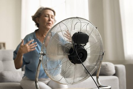 Close up electric ventilator with propeller cooling air blowing to senior older lady suffering from overheating, hypoxia, sitting on sofa with closed eyes, flying hair in blurred background