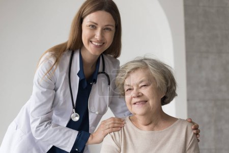 Foto de Happy pretty female doctor with stethoscope embracing older patient, looking at camera, smiling, posing in hospital office, giving medical care, support, assistance. Medical specialist portrait - Imagen libre de derechos