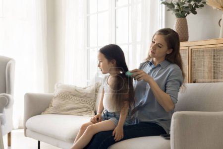 Caring young mom brushing black long hair of cute little girl kid, helping child with girlish beauty care, enjoying childcare, motherhood, maternity leave, family leisure at home