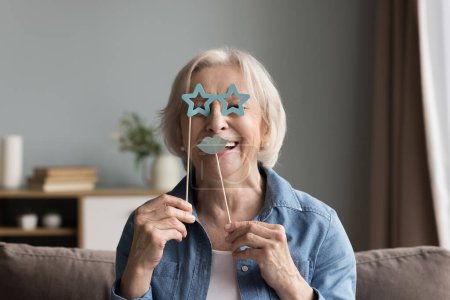 Téléchargez les photos : Joyeux heureux femme âgée appliquant moustache en papier faux et lunettes sur bâton face à face, regardant la caméra, s'amusant, souriant, riant, en utilisant un accessoire pour la prise de photos - en image libre de droit