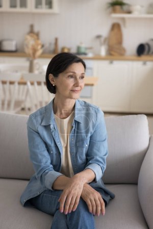 Photo for Thoughtful positive mature senior beautiful woman sitting on home sofa with crossed limbs, looking away, thinking, dreaming, planning future, enjoying calm leisure time at home. Vertical shot - Royalty Free Image