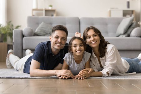 Photo for Happy positive attractive parents and cute tween girl kid resting together on heating floor at home, looking at camera, smiling. Caring mom, dad and daughter lying on bellies, posing for portrait - Royalty Free Image