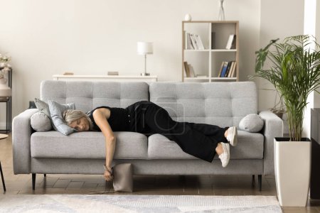 Mujer madura cansada y somnolienta con ropa y zapatos casuales sosteniendo el bolso de mano, descansando en el sofá, acostada sobre el vientre con los ojos cerrados, durmiendo, descansando, haciendo una pausa, sintiendo fatiga, agotamiento