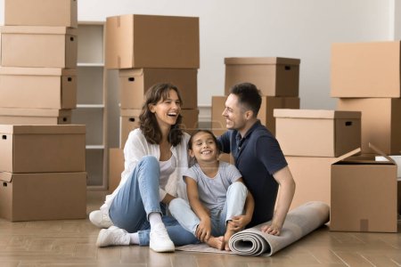 Photo for Cheerful family couple and little kid moving into new apartment after mortgage, real estate property buying, sitting on floor at home with stacked boxes in background, laughing, smiling - Royalty Free Image