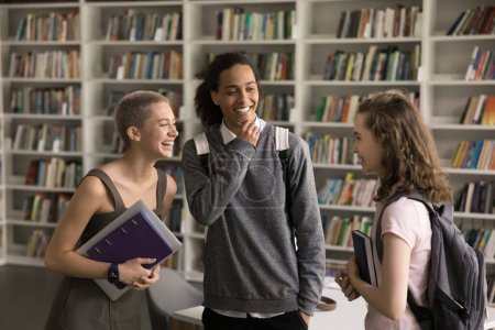 Photo for Cheerful diverse student girls and guy talking in college library together, laughing, discussing education, enjoying friendship. Happy high school classmates with backpacks having fun in campus - Royalty Free Image