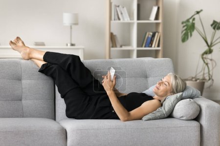 Photo for Lazy relaxed mature woman lying on sofa with smartphone at home, watching online media content, texting message, typing, chatting on Internet, enjoying relaxation with wireless technology - Royalty Free Image