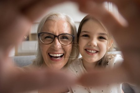 Photo for Excited senior grandma and positive granddaughter girl making finger heart, showing symbol of love, affection, family bonding, looking at camera through hand frame, smiling for close up shot - Royalty Free Image