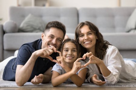 Photo for Cheerful attractive mom, dad and sweet daughter kid joining fingers, showing hand heart shapes, resting together on warm floor at cozy home, looking at camera with toothy smiles for family portrait - Royalty Free Image