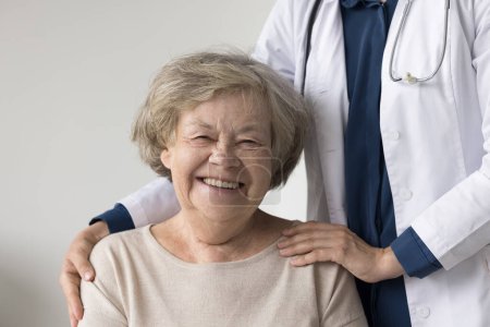 Photo for Portrait of happy positive elderly patient woman posing near geriatrician doctor, looking at camera, smiling, laughing. Cropped shot of practitioner touching shoulders of senior lady - Royalty Free Image