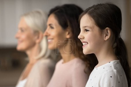 Photo for Happy pretty little tween girl standing by mother and grandma in line, looking away, smiling, laughing, posing for family side portrait. Three female family generations home shot - Royalty Free Image