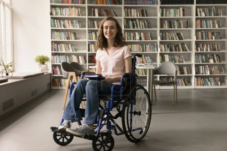 Joven estudiante universitario alegre con discapacidad, visitando la biblioteca del campus, sosteniendo el libro, mirando a la cámara con una sonrisa dentada, promoviendo un ambiente educativo inclusivo