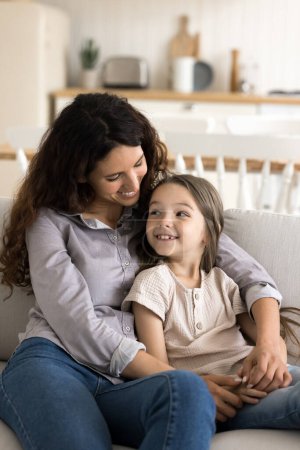 Vertical shot Hispanic woman sit on sofa with pretty daughter, small child enjoy pleasant time with affectionate mommy rest together on couch in cozy flat, smile, lead conversation on weekend at home