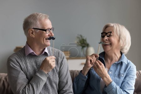 Téléchargez les photos : Heureux couple âgé joyeux de grands-parents jouant avec des accessoires de tir photo, appliquant de fausses moustaches et des lunettes sur les visages, s'amusant, souriant, riant - en image libre de droit