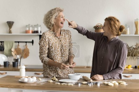 Joyful excited adult daughter woman leaving floury spot on happy mothers face, laughing, having fun at kitchen table with flour, mild, eggs, helping mom to bake cookies, bakery dessert snacks