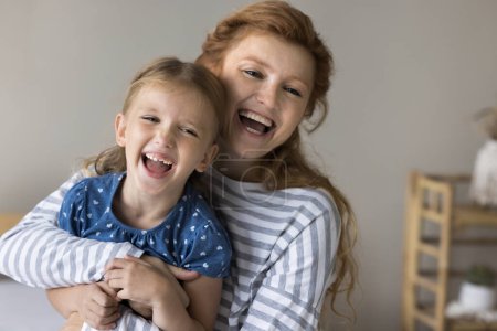 Cheerful red haired mom cuddling, tickling cute kid on sofa, playing with girl, holding shouting joyful child in arms, laughing, enjoying motherhood, family leisure