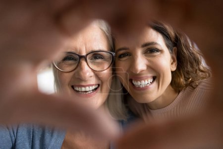 Photo for Cheerful pretty senior mom and adult daughter looking at camera through hand heart, making finger frame, smiling with white healthy teeth, expressing love, family affection, care. Close up portrait - Royalty Free Image