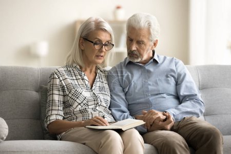 Photo for Caring senior wife reading book aloud to older husband, reviewing written notes in notebook. Elderly retired couple enjoying leisure, literature at home together, holding hands - Royalty Free Image