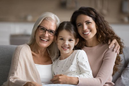 Photo for Happy mom and elderly grandma hugging cute tween girl, posing for family portrait on home couch, looking at camera with toothy smiles, enjoying family bonding, leisure together - Royalty Free Image