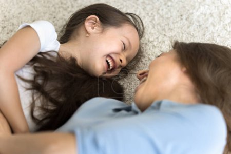 Photo for Happy excited mom and kid relaxing together on soft warm carpeted floor, lying on backs, laughing, having fun, playing. Young mother enjoying family leisure, playtime with girl, motherhood - Royalty Free Image