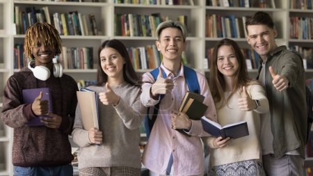 Foto de Happy multiethnic first year college students posing in university library, looking at camera, standing at bookshelves, showing like thumb up gestures, smiling, laughing, recommending education - Imagen libre de derechos