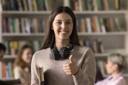 Photo for Cheerful pretty teenage Hispanic student girl showing like hand sign at camera, making thumb up gesture, giving positive feedback, recommending higher education, studying in library. Female portrait - Royalty Free Image