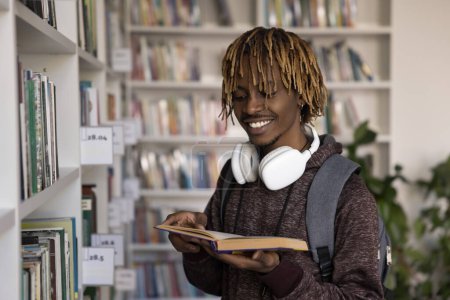 Téléchargez les photos : Heureux étudiant africain dans la bibliothèque universitaire, debout sur les étagères de la bibliothèque, profiter des études, passe-temps de la littérature, obtenir des connaissances, lire livre ouvert, sourire, promouvoir l'éducation collégiale - en image libre de droit