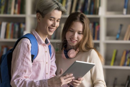 Two cheerful diverse couple of classmates standing together, watching online content on digital gadget. Happy student guy showing presentation on tablet to girlfriend