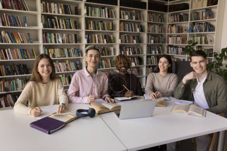 Photo for Cheerful multiethnic teenage students studying together in college library, sitting at table with books, laptop, writing notes, looking at camera, smiling, promoting higher education. Group portrait - Royalty Free Image