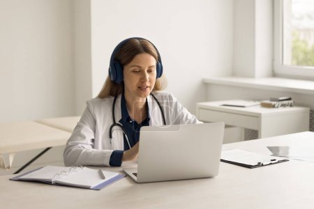 Photo for Focused 35s female doctor in white uniform sits at desk in clinic office in headphones, use laptop gives virtual consultation, talk to patients remotely. On-line counselling, telemedicine, e-health - Royalty Free Image