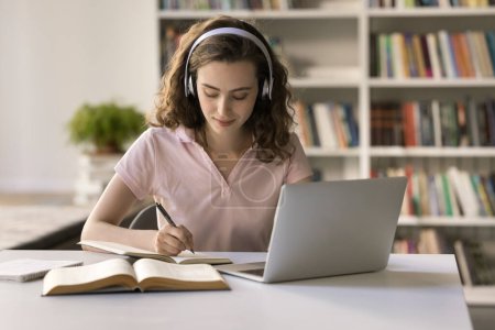 Attraktive Teenager-Studentinnen tragen Kopfhörer, wenn sie in der Bibliothek lernen, sitzen am Schreibtisch mit Laptop, machen Aufgaben, schreiben Aufsätze, hören Audio-Kurse, verbessern Fremdsprachenkenntnisse, lernen im Klassenzimmer