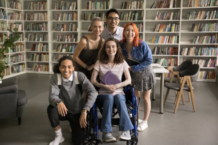 Fünf Studenten posieren in der Universitätsbibliothek für die Kamera. Attraktives Mädchen mit Mobilitätsbehinderung und freundliche Mitschüler, die lächelnd in die Kamera schauen, genießen ihre Freundschaft und ihr Studium
