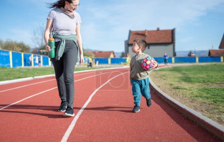 A 3 year old toddler excitedly runs with a soccer ball on a track field with his aunt, indicating an early love for football sports The image captures the innocence and enthusiasm of young sports aspiration