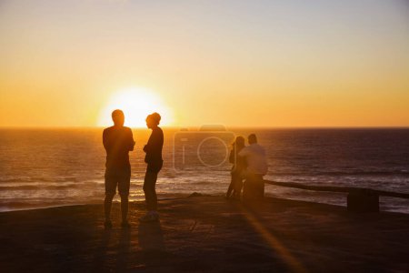 Photo for Silhouettes of the two pairs are on the pier against the background of bright sun and sunset sky. A group of people whose silhouettes are lit by the sunset on the horizon of the ocean. - Royalty Free Image