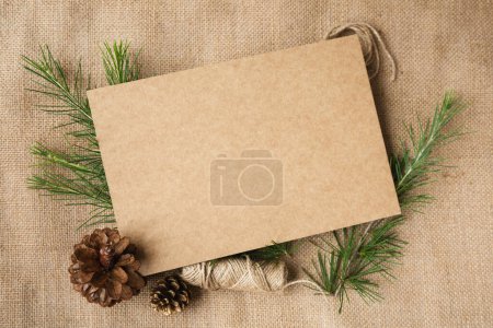 Téléchargez les photos : Une feuille de carton est au milieu de l'image entourée d'une décoration de Noël et de branches et cônes d'épinette sur un fond de toile. Style rétro. Espace de copie. - en image libre de droit