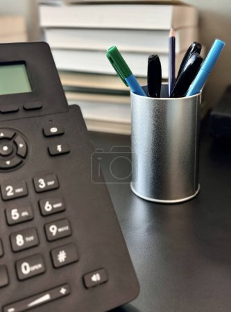  Iron tumbler with pens and pencils, landline black phone is on office desk on background of books in blurred focus.