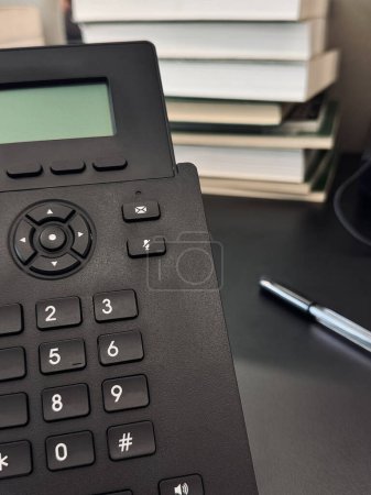 A landline black phone is on an office desk on a background of books in a blurred focus.