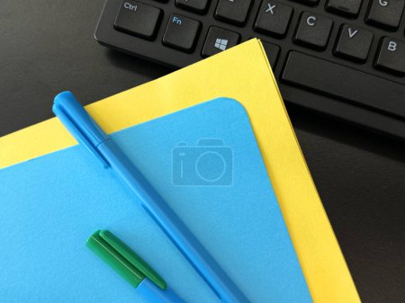 The makers or pens are on blue and yellow paper, and the keyboard in blurred focus are on the office desk.