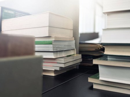 Stacks of books are on an office desk in blurred focus, illuminated by the bright daylight from the window.