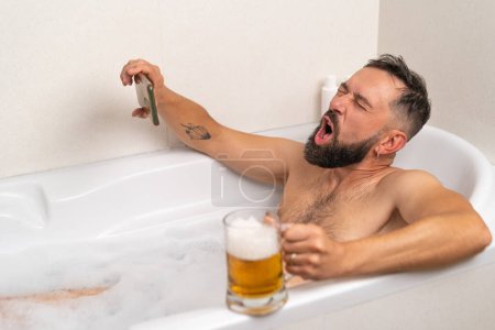 Foto de Hombre barbudo viendo fútbol en el teléfono inteligente mientras bebe una cerveza en el baño con jabones en casa - Imagen libre de derechos