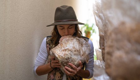 Foto de Happy young woman holding mycelium substrate with lion mane mushrooms and sniffing it in domestic the mushroom farm. - Imagen libre de derechos
