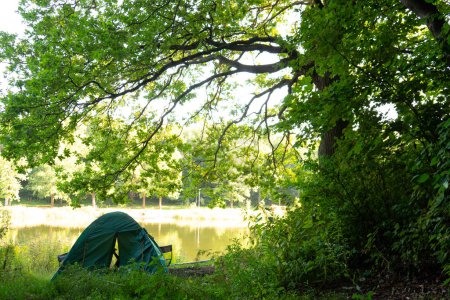 Photo for Picturesque landscape with green camping tent placed under trees branches on shore of river. One shot in series of camping - Royalty Free Image