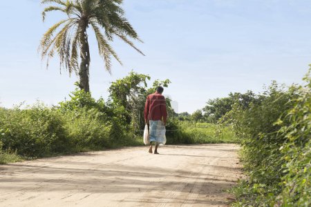 Téléchargez les photos : Un villageois qui va faire du shopping au marché du village. Un lungi portant un homme marchant avec un sac ou un sac dans une journée ensoleillée dans une zone rurale du Bangladesh. - en image libre de droit