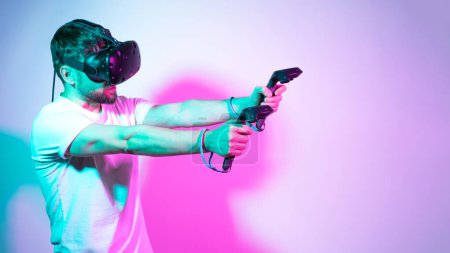 Photo for Bearded man in VR glasses plays in simulation figth and using controllers in in multicolored neon lights colorful background. Concept interactive online game in a virtual world. Copy space, banner. - Royalty Free Image