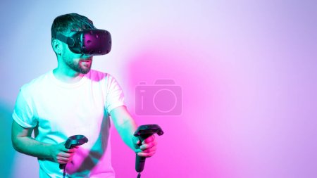 Photo for Concept modern hobby. Bearded millennial man in VR glasses plays in simulation figth and using controllers in gradient neon background. interactive online game in a virtual world. Copy space, banner. - Royalty Free Image