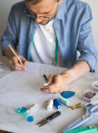 Foto de Un diseñador de bricolaje dibuja un boceto para un nuevo proyecto de vestuario. Una costurera con gafas está trabajando en la mesa. La mano de un diseñador autodidacta con un lápiz. Aficion, estilo de vida, hecho en casa. - Imagen libre de derechos