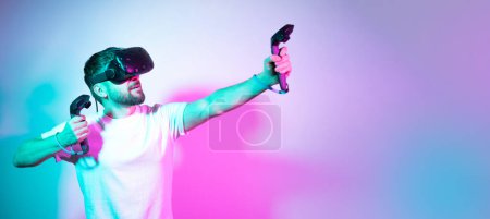 Photo for Concept modern hobby. Bearded millennial man in VR glasses plays in simulation figth and using controllers in gradient neon background. interactive online game in a virtual world. Copy space, banner. - Royalty Free Image
